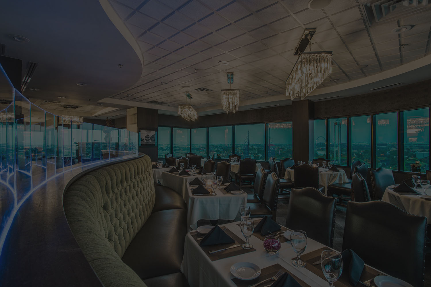The dining room at Mercury Chophouse in Arlington at dusk, city lights beginning to show through the windows behind tables set for two